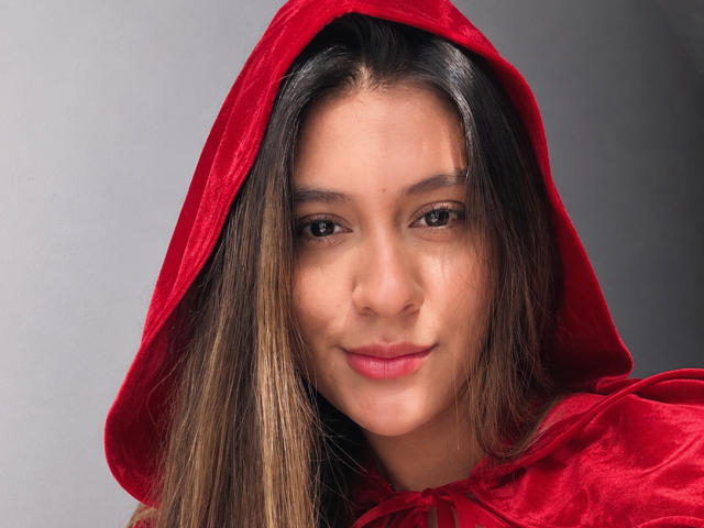 ThaliaAres, avec de longs cheveux bruns, une capuche en velours rouge et un léger sourire, montrant un visage mignon et des yeux marron, portant une cape en velours rouge, regardant droit devant la caméra..