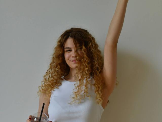 KatherinaHlad with curly blonde hair, white sleeveless top, raising arm, holding drink with black straw, smiling slightly. Her tits are small and her pussy is not visible.