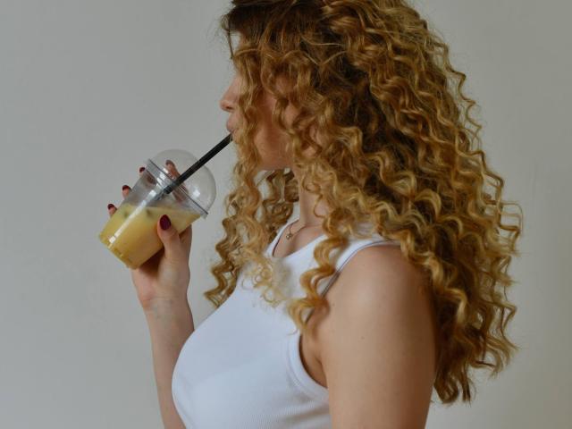 KatherinaHlad with curly blonde hair, white tank top, sipping bubble tea, black straw in her mouth, red nail polish, holding clear cup against plain background.