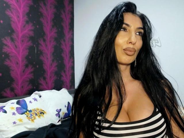 BrunetteBabe69 com cabelos pretos longos, olhos castanhos e seios grandes, vestindo uma blusa listrada, olhando para o lado com lábios curvados, sentada em uma cama com travesseiros coloridos..