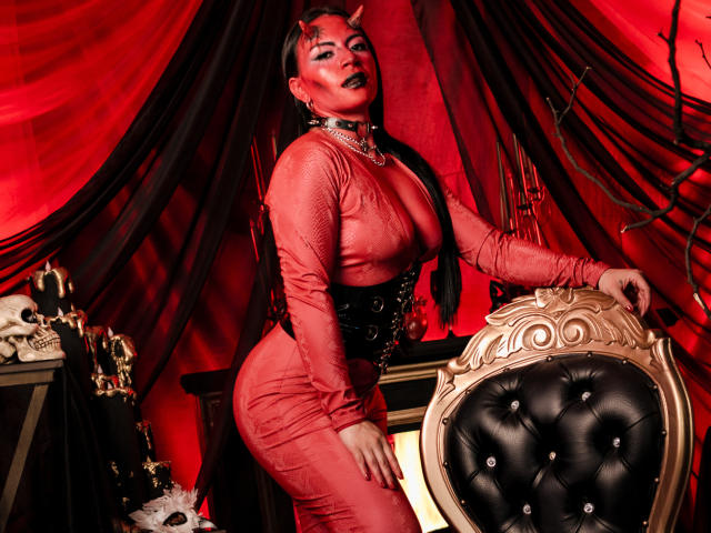 KatalinaJohnson with red skin-tight dress, black corset, and devil horns, showing big tits and round ass, standing by an ornate chair, in a dark, red-lit room.