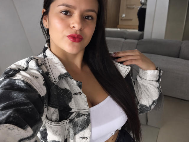 AntonelaBlake with long dark hair, red lipstick, white bra, and black-and-white distressed jacket, showing off her tits and pouty face, hand in hair, in a selfie.