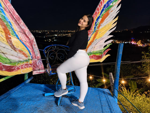 AntonelaBlake met een groot achterwerk, gekleed in witte leggings, een zwarte top en blauwe sneakers, staat op een blauwe stoel met kleurrijke, regenboogkleurige vleugels en kijkt 's nachts terug naar de camera..