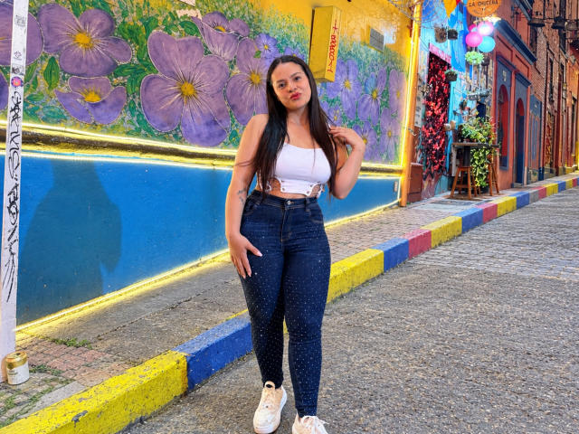 AntonelaBlake with long brown hair, white crop top, showing her tits, and dark blue sparkly jeans, standing on a cobblestone street, hand in hair, looking at camera.