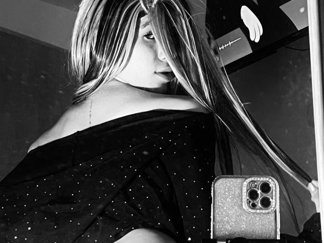 SiaraLatina with long, straight hair, black off-shoulder top, and dark lipstick, holding a glittery phone, looking over her shoulder, showing her bare shoulder and a small tattoo.