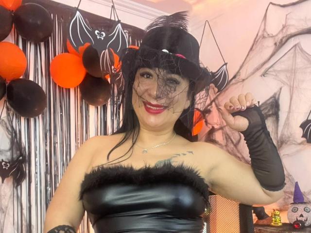 Karimelee in a black leather dress with fur trim, wearing a witch hat, veil, and black gloves, smiling with red lipstick, showing tattoo on chest, and winking.