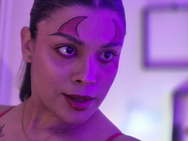 TeylorJenkins has purple makeup on her forehead, red lipstick, and a tattoo on her shoulder. She's looking to the side with a slight smile, under purple lighting.