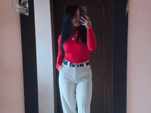 IsaLawrence with long black hair, red long-sleeve top, white pants, black belt, and necklace, taking a mirror selfie, showing her ass and small tits.