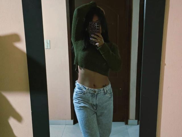 IsaLawrence with dark hair, green sweater, and light blue jeans, showing a slim midriff, taking a mirror selfie, covering her face with a phone.