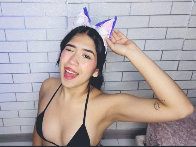 HannaThorne with black bikini top, cat ears, winking, smiling, dark hair, small tits, tattoo on right arm, white brick wall background, pink towel, right arm raised.