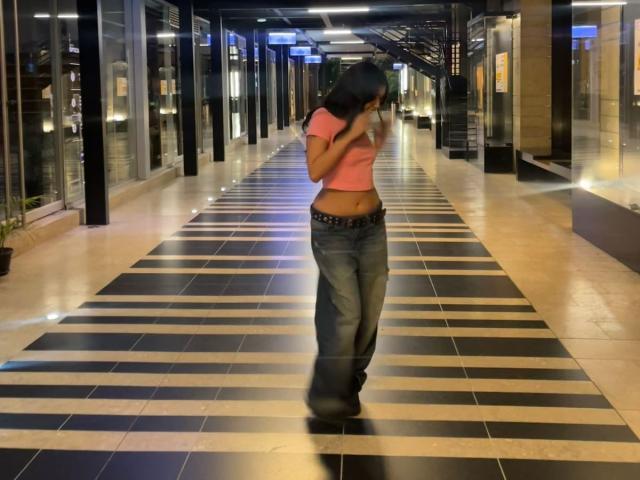 HannaThorne stands in a hallway, covering her face with one hand, wearing a pink crop top, blue jeans, and a black belt, with a visible belly button.