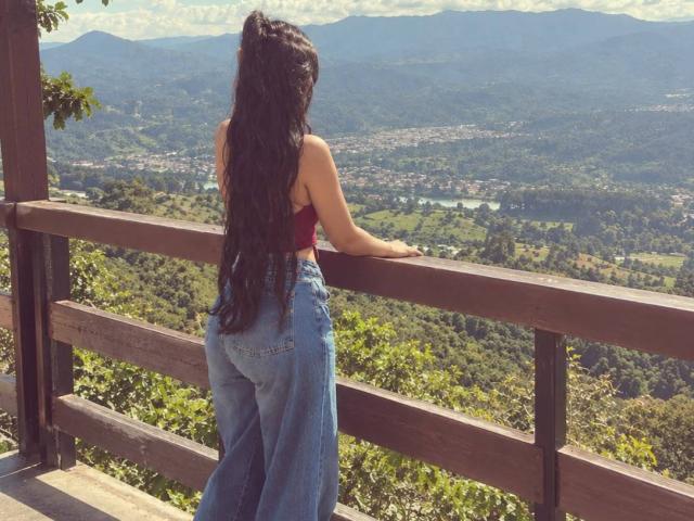 AmyLambert with long black wavy hair, red crop top, and blue high-waisted jeans, standing with her back to a wooden railing, looking at a mountainous valley.