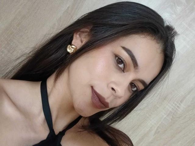 AlanaBlake has long black hair, brown eyes, and dark lipstick. She's wearing a black halter top and a gold heart earring, with a slight smile and smooth skin.