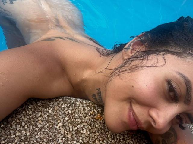 ElectraRoss is lying topless in a pool, her wet brown hair against her shoulder, smiling slightly, with tattoos visible on her neck, and resting on a gravel surface.