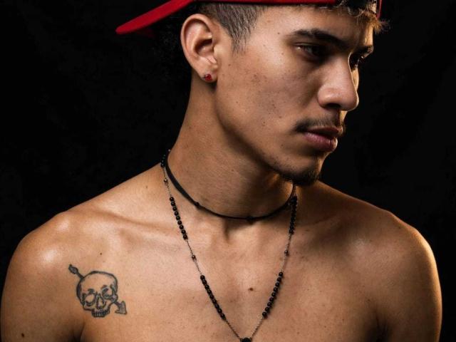SkinnyCuteAlyer with short hair, red cap, black beads necklace, small red earbuds, skull tattoo on left shoulder, looking down, black background, shirtless, brown skin, slight mustache.