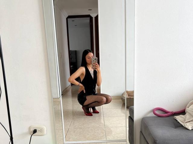 NiaWhitee, squatting in front of a mirror, wearing a black dress, sheer black stockings, and red heels, holding a phone, with visible ass and medium tits.
