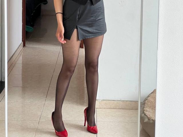 NiaWhitee's legs in black sheer pantyhose, red high heels, and a grey mini skirt with a side slit, standing on a tiled floor, black wristband on her right wrist.
