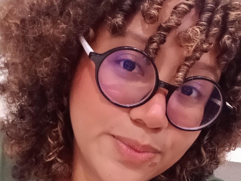 MaraSilk69 with curly brown hair, purple-tinted glasses, and a subtle smile. Her eyes reflect a bit of the purple from the glasses. She's looking directly at the camera.
