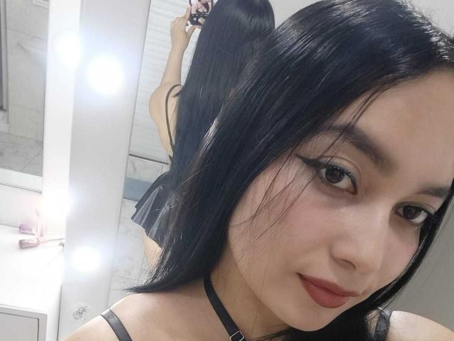 MelaniCoox with black hair, thick eyeliner, red lipstick, black choker, looking at mirror showing her back and black dress, reflecting her ass. Bathroom with lights in mirror.