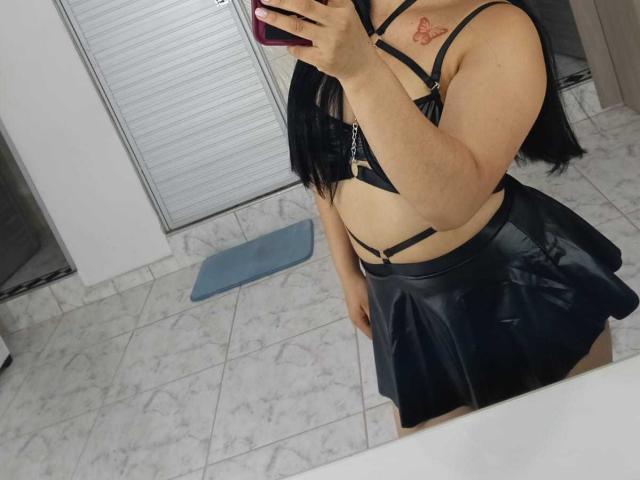 MelaniCoox with black strappy bra and shiny black skirt, holding phone in mirror selfie. Tattoo on right shoulder. Long black hair, white tiled floor, blue mat.