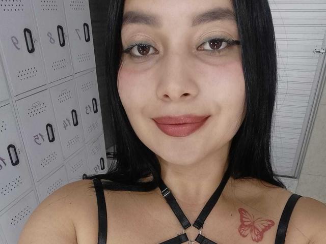 MelaniCoox with black hair, brown eyes, and pink lipstick, wearing a black strappy top, showing a red butterfly tattoo on her chest, smiling at the camera.