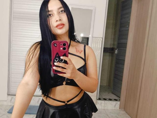 MelaniCoox with long black hair, wearing a black strappy bra and skirt, holding a pink phone, showing a small tattoo on her right shoulder, and a slight smile.