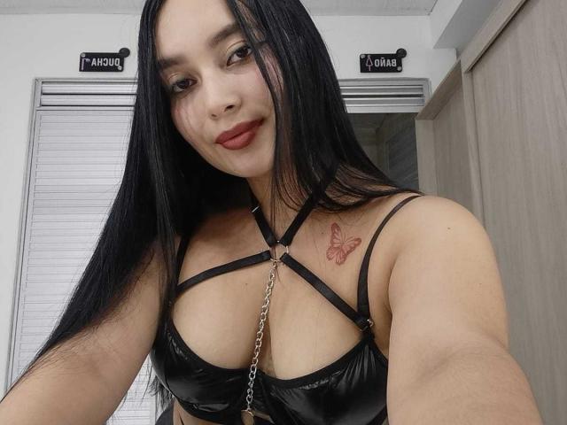 MelaniCoox with long black hair, red lipstick, and a butterfly tattoo on her right shoulder, wearing a black strappy leather top, showing her tits and a silver chain.