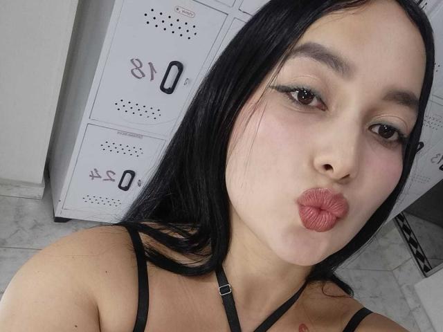 MelaniCoox has black hair, wearing a black strap top, pouting her pink lips, with a tattoo on her left shoulder, brown eyes, and a light skin tone.