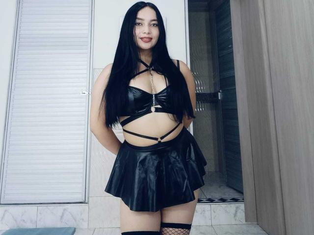 MelaniCoox with long black hair, wearing a black leather strappy top, black leather mini skirt, and fishnet thigh-highs, standing in a room with a wooden door.