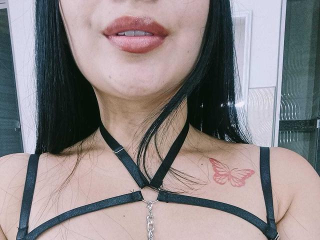 MelaniCoox with black strappy lingerie, red butterfly tattoo on right shoulder, glossy pink lips, and long black hair. Her tits are partially visible, and she's wearing a metal chain.