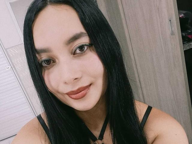 MelaniCoox has long black hair, brown eyes, and a slight smile with pink lipstick. She's wearing a black strap top, showing her small tits. Her skin is light.