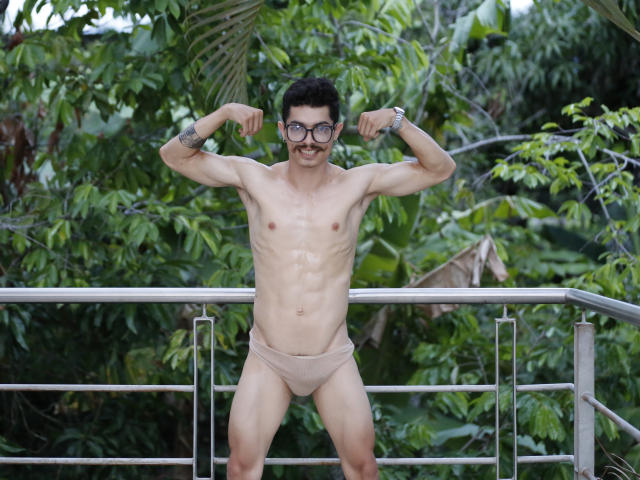 TaylorTele, skinny dude with glasses, small tits, wearing beige briefs, flexing biceps, mustache, tattoos on forearms, standing in front of metal railing, surrounded by greenery.