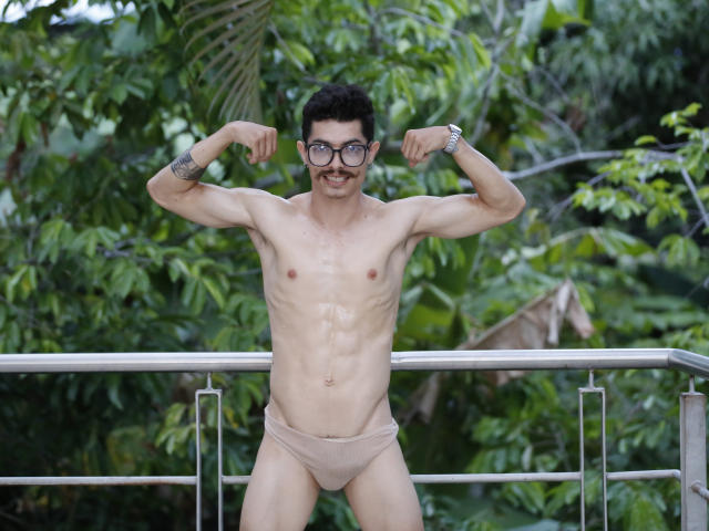 TaylorTele, a skinny dude with glasses, flexes his arms, showing off his small, flaccid ass and tiny, uncut cock in beige briefs, standing in front of greenery.