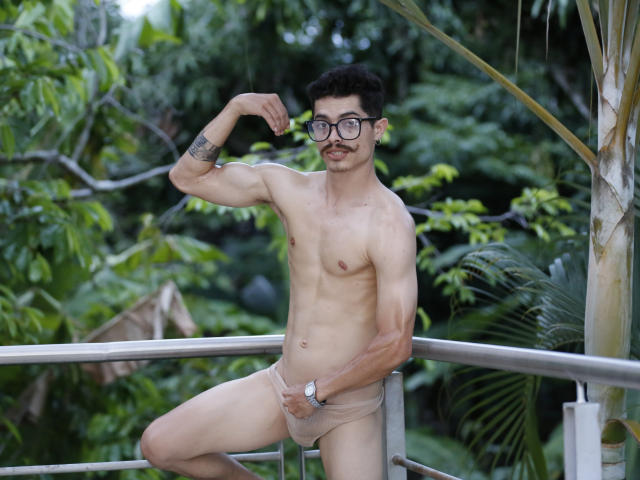 TaylorTele, a slim, muscular dude with a mustache, glasses, and a tattoo, flexes his bicep while wearing beige jockstrap, standing on a metal railing, outside.