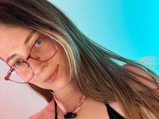AmberWoody with long brown hair, pink glasses, and a subtle smile, wearing a pink necklace and black top, has cute tits and a sexy, fresh-faced look.