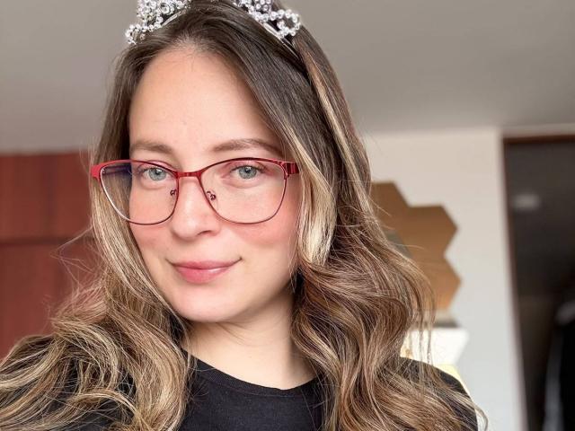 AmberWoody with wavy blonde hair, red glasses, and a silver tiara, smiling softly, showing her cute ass cheeks through a black top. Her tits are small and cute.
