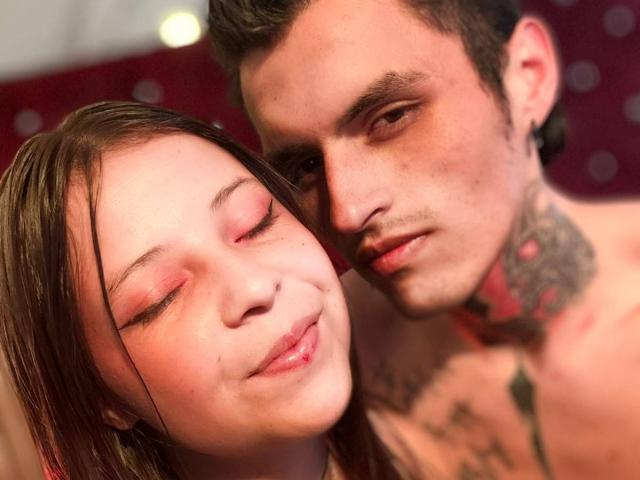 GraceAndTony with pink eye makeup, nose piercing, and brown hair leans on Tony, who has tattoos and black ear plugs, with a slight smile. Tony's face is close to hers.