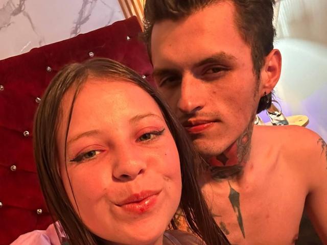 GraceAndTony with puffy red lips, brown hair, and mascara, next to Tony with tattoos, ear gauges, and a shirtless upper body. Both are smiling in a warm, orange-lit room.