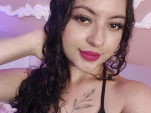 MiiaBennet with curly black hair, pink lipstick, and a tattoo on her chest, wearing a black halter top, smiling at the camera. Her tits are partially visible.