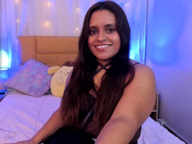 ValerieSynclair with long brown hair, smiling, wearing a black top with a criss-cross strap, sitting on a bed with a fluffy pillow and stuffed animal.