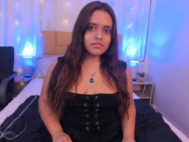 ValerieSynclair, with long brown hair, wearing a black corset, sitting on a bed, has medium tits, and a necklace with a blue pendant, in a blue-lit room.