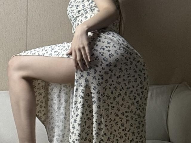 SandiWimbs' white floral dress, showing her right leg raised, hand on her thigh, exposing her bare leg, her ass slightly visible, in a softly lit indoor setting.