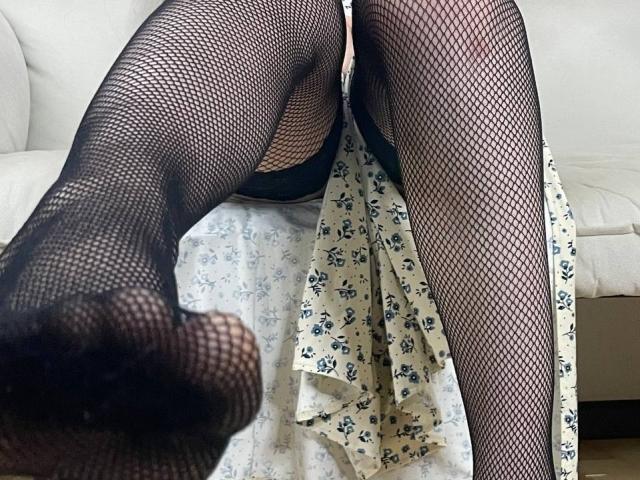 SandiWimbs' legs in black fishnet stockings, feet up, showing her ass and pussy, with a floral skirt bunched up, on a white couch.