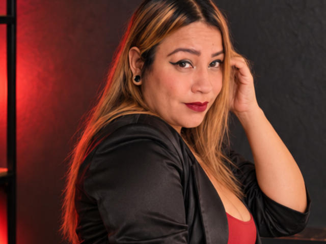 KinkyHouseWife with blonde hair, black earrings, red lipstick, black satin jacket, and red top, looking over shoulder, hand in hair, red light in background, slightly smiling.