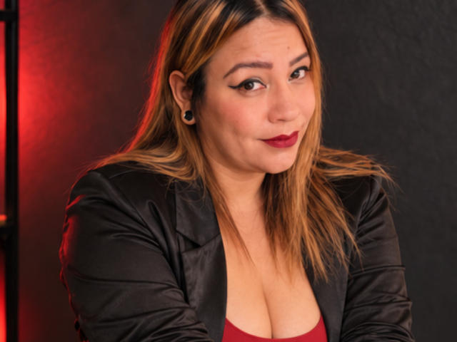 KinkyHouseWife with medium tits, wearing a black leather jacket, red top, and dark red lipstick, has blonde hair, black ear gauges, and a slight smile in a photo.
