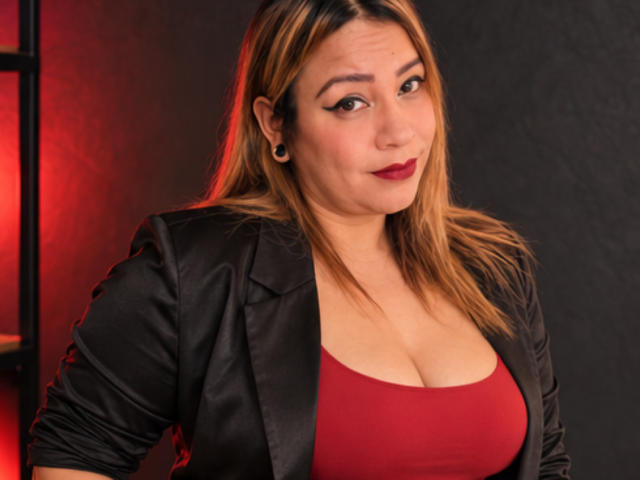 KinkyHouseWife has blonde hair, black leather jacket, red tank top showing her big tits, and black ear gauges, with red lighting in the background. She's smiling slightly.
