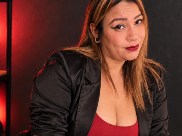 KinkyHouseWife with blonde hair, black leather jacket, red top, showing her tits, dark red lipstick, black ear gauges, smiling slightly, red light on the left.