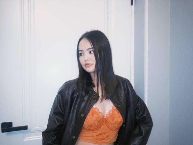 Sovelle with black hair, wearing an orange lace bra and black leather jacket, standing in front of a white door. Boobs are visible under the lace.