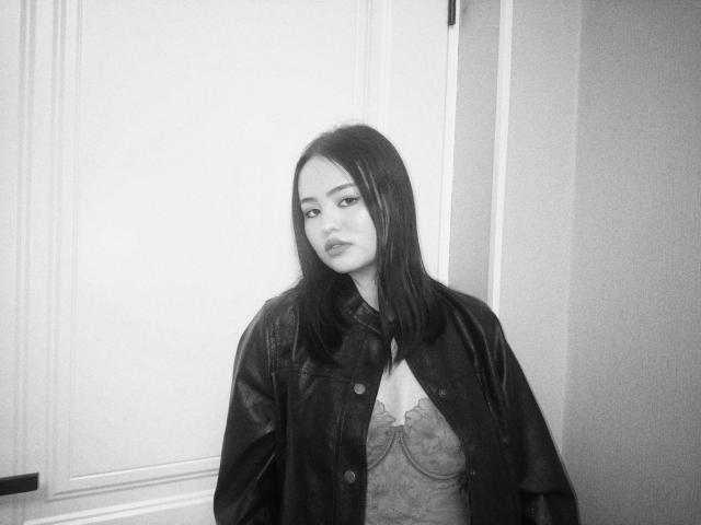 Sovelle has dark, straight hair, wearing a leather jacket over a lace-up bralette, looking off to the side with a serious expression in a black-and-white photo.