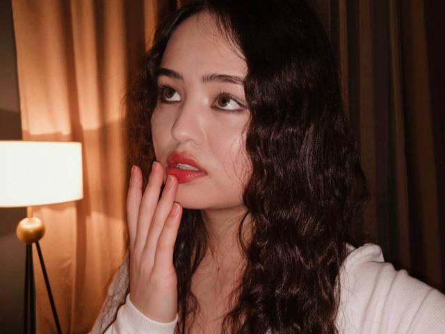 Sovelle with curly black hair, red lips, and white top, hand touching face, looking to the side, warm light from lamp on left, soft brown curtains in background.